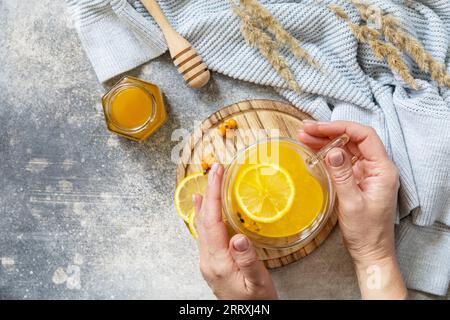 Relaxation et phytothérapie. Thé chaud à base de vitamine d'argousier dans une tasse en verre avec des baies fraîches d'argousier, du miel et du citron dans les mains de Banque D'Images