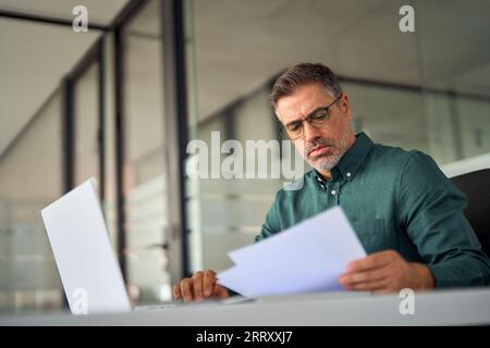 Cadre occupé d'âge moyen tenant des documents à l'aide d'un ordinateur portable au bureau. Banque D'Images
