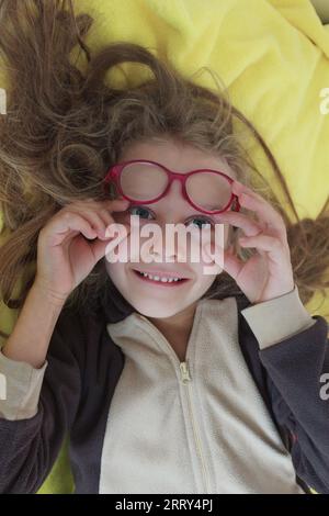 Une petite fille drôle avec des lunettes se trouve sur une couverture jaune sur le lit et regarde dans le cadre. Banque D'Images