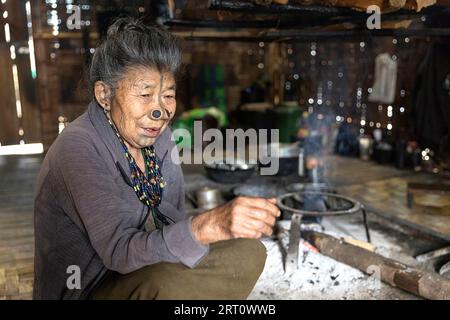 Femme locale de la tribu Apatani avec des tatouages de visage traditionnels se trouvant près de la cheminée dans une maison traditionnelle à Ziro dans l'Arunachal Pradesh, Inde Banque D'Images