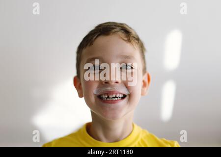 Mignon garçon boit du kéfir ou du lait. Portrait drôle d'un garçon avec une moustache de lait. Banque D'Images