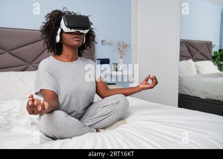 Jeune femme africaine méditant avec des lunettes VR assis sur le lit. Banque D'Images