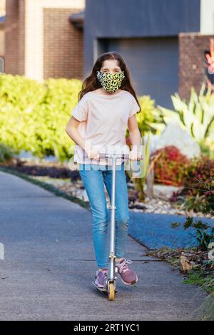 Une jeune fille caucasienne joue avec son scooter dans la rue par une soirée ensoleillée en Australie tout en portant un masque COVID Banque D'Images