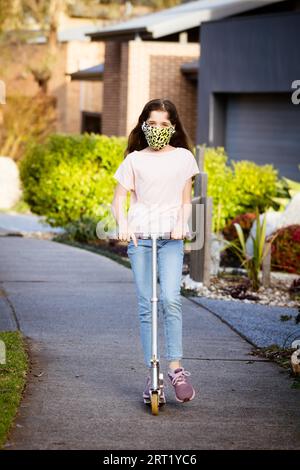 Une jeune fille caucasienne joue avec son scooter dans la rue par une soirée ensoleillée en Australie tout en portant un masque COVID Banque D'Images