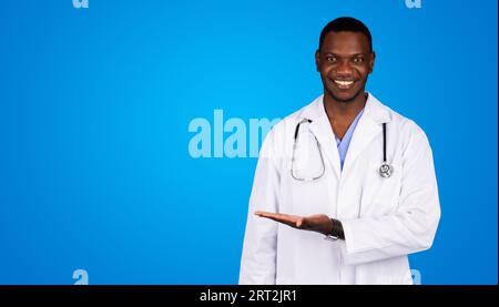 Heureux jeune homme afro-américain docteur en manteau blanc tenant l'espace vide sur la main pour l'annonce et l'offre Banque D'Images