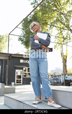 mignonne femme d'affaires senior européenne aux cheveux gris avec une coiffure élégante et des lunettes est habillée dans une veste grise avec un ordinateur portable dans la rue Banque D'Images