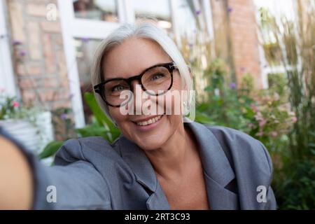 femme rêveuse mature aux cheveux gris habillée pour l'été fait un selfie au téléphone tout en étant assise dans la rue Banque D'Images