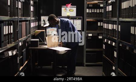 Un enquêteur afro-américain habillé professionnellement examine des preuves confidentielles dans un espace de bureau organisé. Détective privé détenant certains dossiers sur des étagères pour étude et analyse médico-légale. Banque D'Images