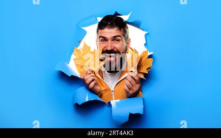 Homme barbu heureux regardant à travers le trou de papier avec des feuilles d'automne. Collection de vêtements d'automne. Homme souriant dans des vêtements chauds avec des feuilles d'érable. Automne Banque D'Images