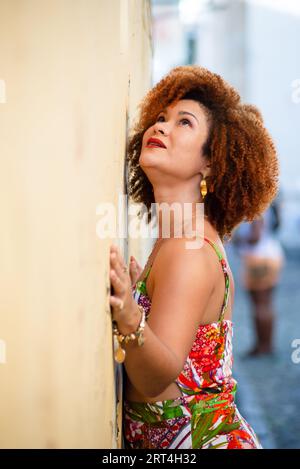 Belle femme aux cheveux roux avec son visage et son corps appuyé contre un vieux mur de couleur claire d'une rue. Personne heureuse voyageant. Banque D'Images