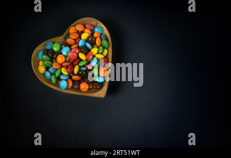 pile de bonbons au chocolat colorés dans un bol en bois en forme de coeur sur fond noir, vue de dessus Banque D'Images