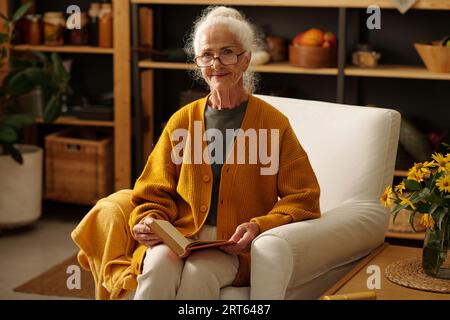 Heureuse femme âgée avec le livre ouvert regardant la caméra tout en étant assis dans un fauteuil par une petite table en bois avec bouquet de fleurs sauvages jaunes Banque D'Images