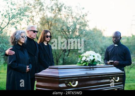 Jeune pasteur avec Sainte Bible et famille de trois debout par cercueil couvert avec des fleurs blanches sur le dessus pendant le service funéraire sur le cimetière Banque D'Images