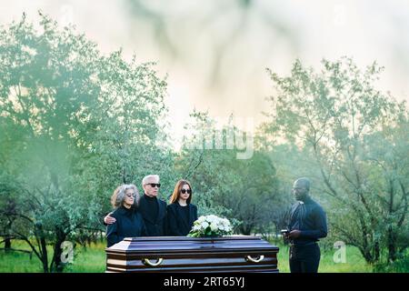 Long plan de deuil famille de trois et prêtre en tenue noire entourant cercueil en bois avec des fleurs blanches sur le dessus au service funéraire Banque D'Images