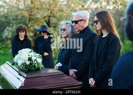 Famille de trois personnes en deuil portant des lunettes de soleil et des vêtements de deuil debout devant un cercueil avec des fleurs blanches sur le couvercle pendant le service funéraire Banque D'Images