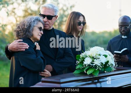 Famille de deuil de trois et prêtre en tenue noire debout devant un cercueil avec des fleurs blanches sur le dessus pendant les funérailles au cimetière Banque D'Images