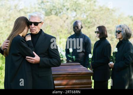 Homme mûr en deuil en costume noir embrassant sa fille en deuil tout en exprimant sa sympathie pour la perte de cher ami ou membre de la famille Banque D'Images