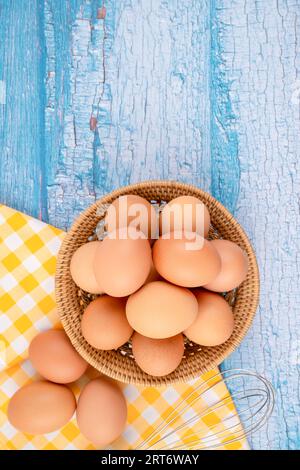 Vue aérienne des œufs de poule bruns dans le panier de tissage et un fouet sur fond en bois bleu. Banque D'Images