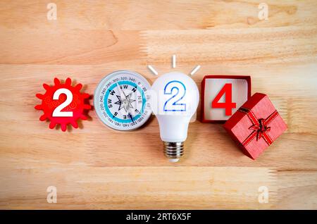 Bonne année 2024. numéro de 2024 ans avec des éléments d'affaires, ampoule créative, boussole, engrenage rouge et boîte-cadeau ouverte sur fond de table en bois. Banque D'Images
