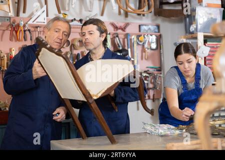 Portrait de trois restaurateurs menuisiers rénovant une chaise victorienne et un cadre de miroir en atelier Banque D'Images