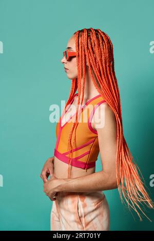 Vue latérale de jeune femme confiante avec de longues tresses afro orange portant des lunettes de soleil et un haut orange regardant loin sur fond turquoise Banque D'Images