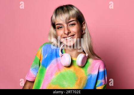 Jeune femme positive avec des piercings de visage et des cheveux blonds et des écouteurs autour du cou dans un t-shirt coloré debout sur fond rose Banque D'Images