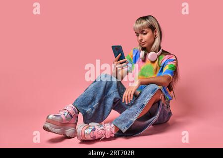 Corps entier de jeune femme en t-shirt coloré et jeans avec des écouteurs autour du cou assis sur le sol en utilisant un téléphone portable sur fond rose Banque D'Images