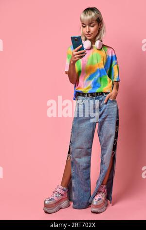 Corps complet de jeune femme en t-shirt coloré et jeans avec écouteurs autour du cou debout à l'aide d'un téléphone portable sur fond rose Banque D'Images