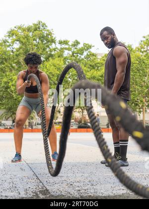 Athlète féminine afro-américaine forte s'exerçant avec des cordes de combat sur un terrain de sport avec instructeur masculin personnel pendant l'entraînement Banque D'Images