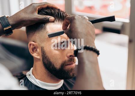 Angle élevé de la récolte anonyme afro-américain barbier utilisant le rasoir tout en donnant la coupe de cheveux à l'homme barbu brutal dans le salon de coiffure moderne Banque D'Images