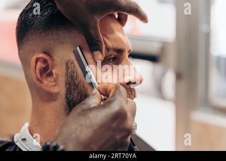 Angle élevé de la récolte anonyme afro-américain barbier utilisant le rasoir tout en donnant la coupe de cheveux à l'homme barbu brutal dans le salon de coiffure moderne Banque D'Images