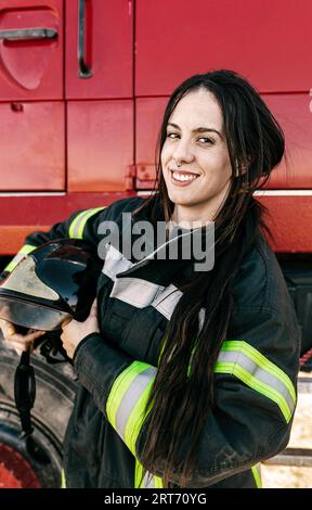 Jeune pompier féminin positif avec queue de cochon noire tenant le casque tout en portant l'uniforme debout contre le moteur de pompiers rouge Banque D'Images