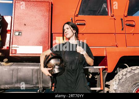 Jeune pompier féminin avec queue de cochon noire tenant le casque tout en portant l'uniforme debout contre le moteur de pompiers rouge Banque D'Images