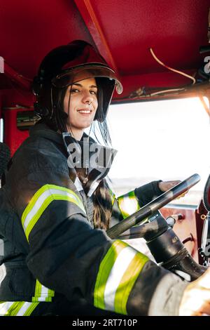 Jeune femme pompier positif en uniforme de protection et casque conduisant un moteur de pompiers rouge tout en travaillant au service d'urgence regardant la caméra Banque D'Images