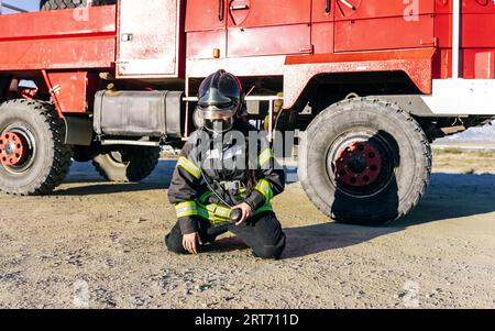 Pompier féminin anonyme portant un uniforme et un masque de protection assis près d'une camionnette de pompiers sous le ciel bleu en journée ensoleillée Banque D'Images