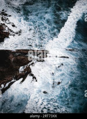 Photo aérienne verticale de l'océan fort et puissant avec d'énormes vagues. Rochers et une partie de falaise à l'intérieur de l'océan bleu orageux - vue de dessus. ph sombre et moelleux Banque D'Images