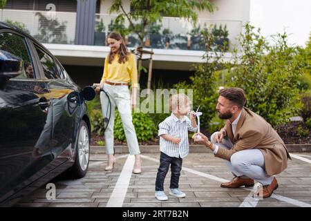 Portriat de femme chargeant la voiture électrique dans la rue, tandis que mari et fils attendent. Banque D'Images