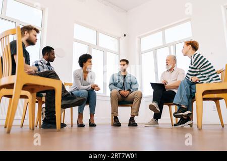 Vue à faible angle de psychologue coach masculin mature confiant parlant assis en cercle pendant la thérapie de groupe avec des personnes d'âges divers et différents Banque D'Images