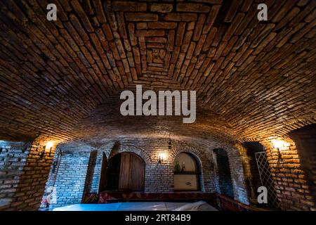 Une voûte en tonneau faite de briques rouges dans un bâtiment patricien historique à Tbilissi : la voûte enjambe un haut hall, avec des appliques murales et un passage en arc de bois le long des murs. Cette technique de construction – briques sans armature en acier, maintenues en place par la géométrie et une clé de voûte – a été largement répandue dans l’architecture caucasienne jusqu’au XIXe siècle. De telles constructions sont maçonnées sur une ossature vide et ne sont stables que grâce à la clé de voûte au point le plus élevé. Anton Chekhov Street, Zemo Avlabari, Metromsheni, Isani District, Tbilissi, 0144, Géorgie Banque D'Images