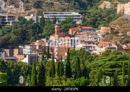 Le quartier des bains d'Abanotubani (géorgien : აბანოთუბანი) baigné par la lumière du matin sur le flanc en contrebas de la forteresse de Narikala dans la vieille ville de Tbilissi, en Géorgie. Le minaret en brique de la mosquée Juma s'élève parmi les maisons aux toits de tuiles rouges. En bas à gauche, un arc pointu carrelé s'ouvre, probablement la façade du bain Orbeliani (Chreli Abano, "bain coloré"). De petites dômes bleus se courbent à côté. En haut à droite, les murs et une tour de la forteresse se dressent sur le rocher. Un long bâtiment moderne et une coquille de bâtiment se trouvent plus haut sur le flanc. Des cyprès et des arbres caduques poussent au premier plan. La ville n Banque D'Images