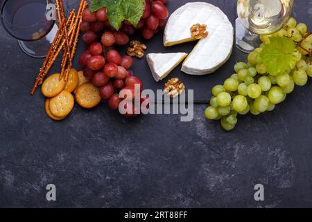 Planche d'ardoise avec brie au fromage, raisins blancs et rouges, crackers noix et vin sur pierre noire, vue aérienne horizontale Banque D'Images