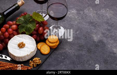 Planche d'ardoise avec brie au fromage, raisins, crackers noix et vin sur pierre noire, vue aérienne horizontale Banque D'Images