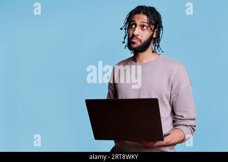 Homme arabe pensif pensant au rapport d'affaires tout en tenant l'ordinateur portable et en regardant vers le haut. Personne réfléchie avec expression faciale intriguée analysant le projet de démarrage sur ordinateur portable Banque D'Images