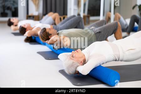 Active senior femme effectue des exercices avec un rouleau pilates tout en étant allongé sur un tapis dans le hall d'un studio de fitness moderne. Banque D'Images