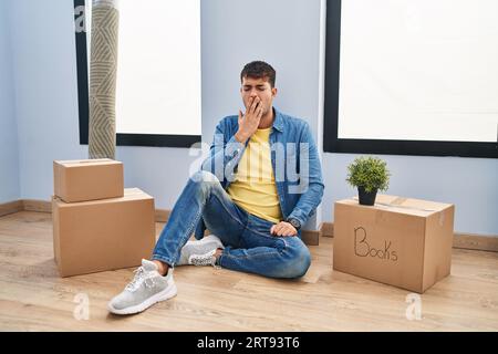 Jeune homme hispanique assis sur le sol à la nouvelle maison ennuyé bâillant fatigué couvrant la bouche avec la main. agité et somnolence. Banque D'Images