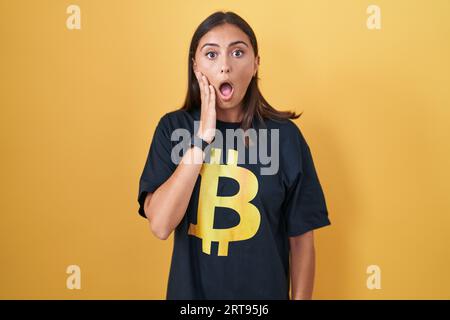 Jeune femme hispanique portant un t-shirt bitcoin effrayé et choqué, surprise et expression stupéfaite avec les mains sur le visage Banque D'Images