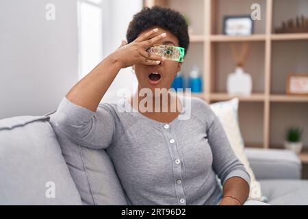 Jeune femme afro-américaine portant des lunettes de réalité virtuelle assise sur le canapé regardant dans le choc couvrant le visage et les yeux avec la main, regardant à travers fi Banque D'Images