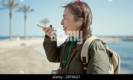 Femme hispanique mature avec cheveux gris touriste portant sac à dos parlant au téléphone au bord de la mer Banque D'Images