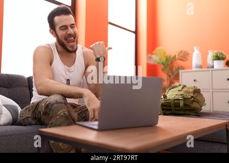 Jeune homme hispanique portant l'uniforme de l'armée de camouflage faisant appel vidéo à la maison pointant le pouce vers le côté souriant heureux avec la bouche ouverte Banque D'Images