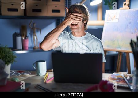 Homme d'âge moyen assis au studio d'art avec ordinateur portable la nuit souriant et riant avec la main sur le visage couvrant les yeux pour la surprise. concept aveugle. Banque D'Images